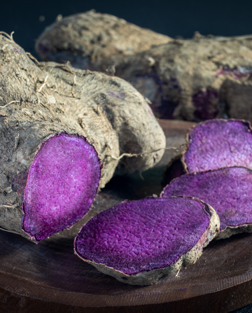 Lila Süßkartoffeln liegen auf einem Holzbrett, einige sind ganz, andere in Scheiben geschnitten. Die Schnittflächen zeigen eine leuchtend lila Farbe. Der Hintergrund ist dunkel.