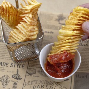 Knickrige Kartoffelchips in einem Korb, daneben eine Schüssel mit roter Sauce. Eine Hand hält einen langen Stapel Chips, der in die Sauce getaucht wird. Der Hintergrund zeigt eine bedruckte Unterlage.