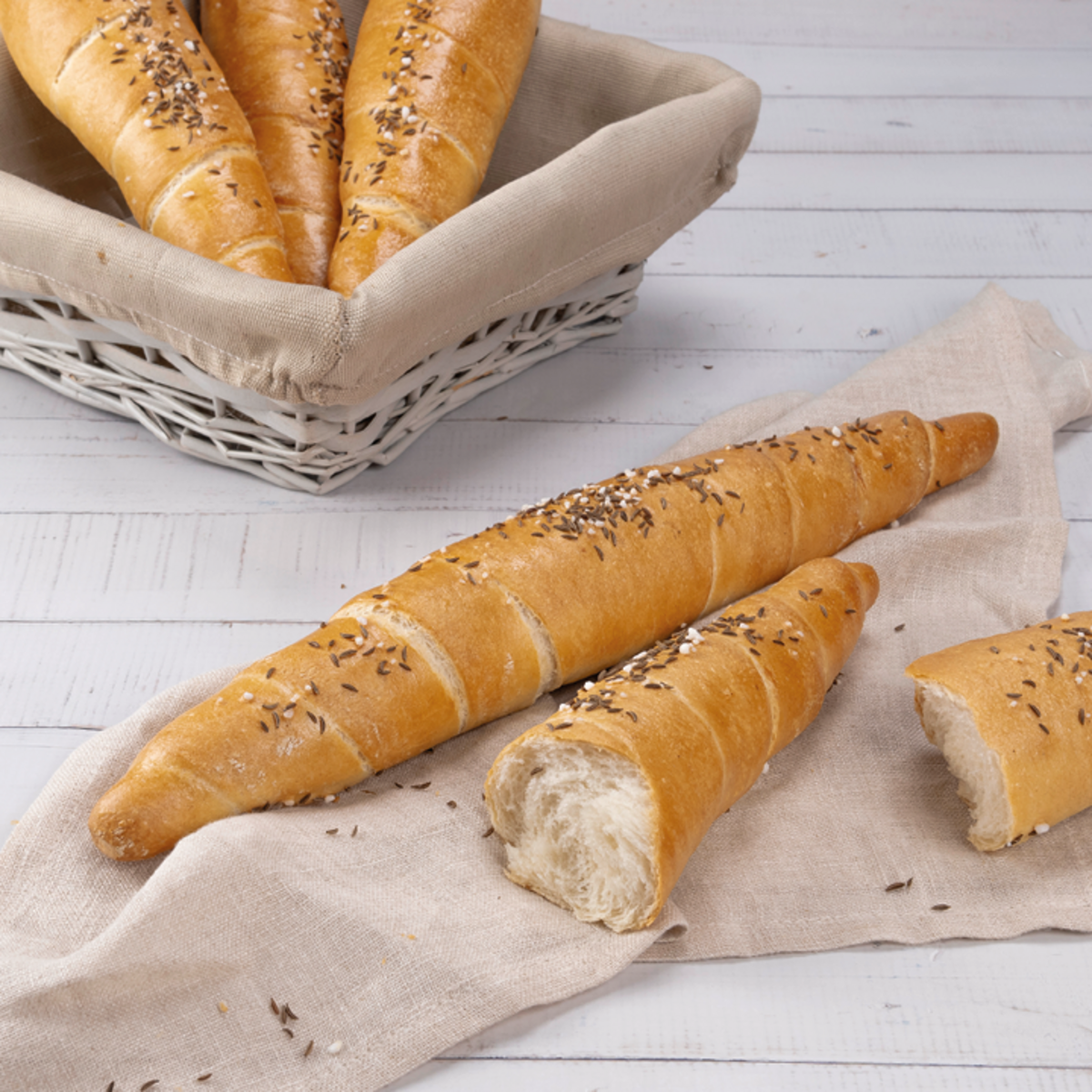 Lange, gebogene Brötchen mit Kümmel und Salz, teilweise auf einem Leinentuch und in einem Korb angeordnet. Einige Brötchen sind aufgeschnitten, das Innere ist weich und hell.