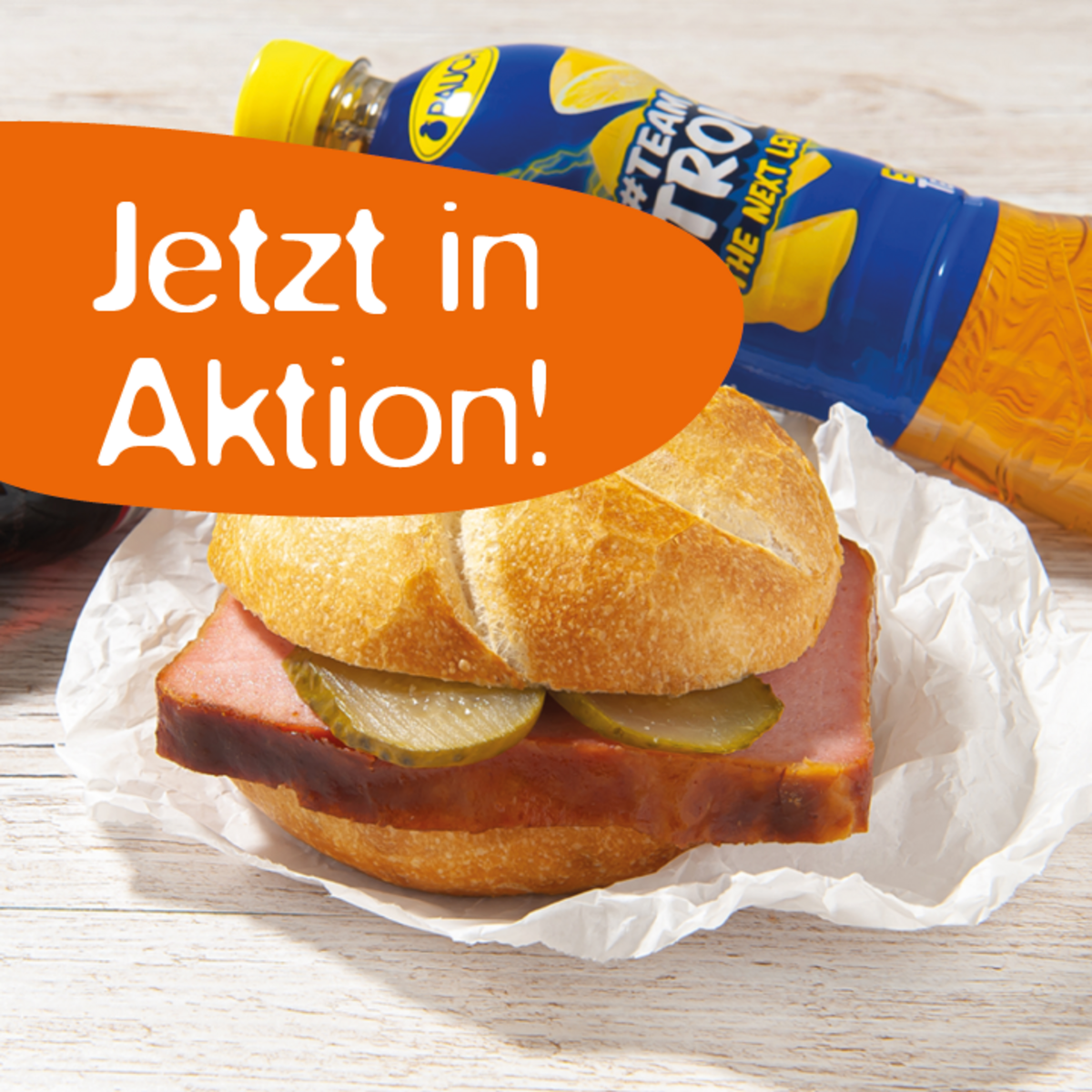 Ein Sandwich mit Schinken und Gurkenscheiben auf einem Stück Papier, daneben eine Flasche Limonade mit gelbem Etikett. Ein orangefarbener Text sagt "Jetzt in Aktion!