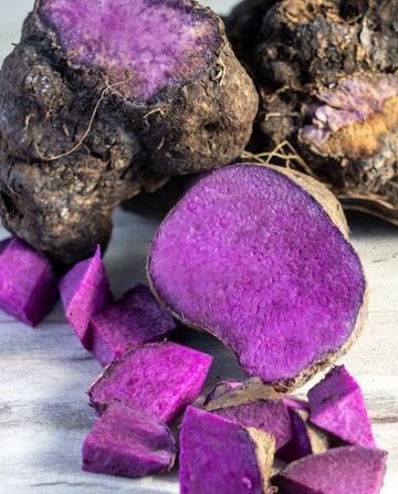 Lila Süßkartoffeln, einige ganz und einige in Würfeln geschnitten, liegen auf einer hellen Oberfläche. Die Schnittflächen zeigen die leuchtend lila Farbe.