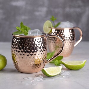 Zwei kupferne Becher mit Eiswürfeln und frischer Minze, umgeben von Limettenhälften und einer ganzen Limette auf einem hellen Tisch.