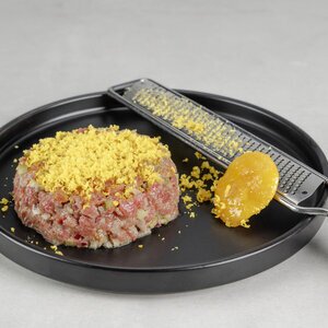 Rohes Hackfleisch auf einem schwarzen Teller, bedeckt mit gelben Bröseln. Neben dem Fleisch liegt eine Reibe mit weiteren Bröseln und einer Portion gelber Paste.