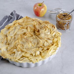 Ein runder, gebackener Teig mit goldbrauner Oberfläche liegt auf einem grauen Tisch. Daneben steht ein Glas mit einer süßen Füllung und ein roter Apfel. Ein gestreifter Küchentuch ist ebenfalls sichtbar.