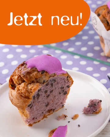 Ein Stück Muffin mit lila Zuckerguss auf einem weißen Teller, daneben ein ganzer Muffin auf einem Glasuntersetzer. Im Hintergrund ein lila Tischset mit weißen Punkten. Text: "Jetzt neu!