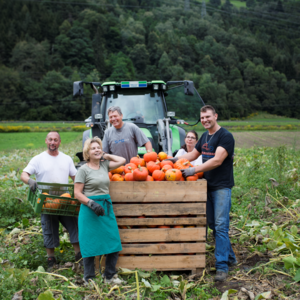 Fünf Personen stehen auf einem Feld und lächeln. Sie halten eine Holzkiste voller Kürbisse. Im Hintergrund ist ein Traktor und bewaldete Hügel zu sehen.