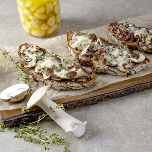 Drei belegte Brote mit Pilzen und Käse auf einem Holzbrett, umgeben von frischen Pilzen und Thymian. Im Hintergrund ein Glas mit eingelegten Zwiebeln.
