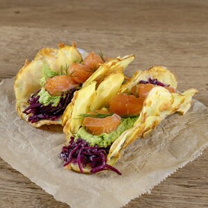 Drei knusprige Tacos aus Kartoffeln, gefüllt mit Avocado, Rotkohl und Lachs, garniert mit frischem Dill, auf braunem Papier platziert.