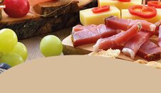 Auf einem Tisch liegen verschiedene Lebensmittel: Scheiben von Schinken, Käsewürfel, Trauben und Brot. Einige rote Paprikastücke sind ebenfalls sichtbar.