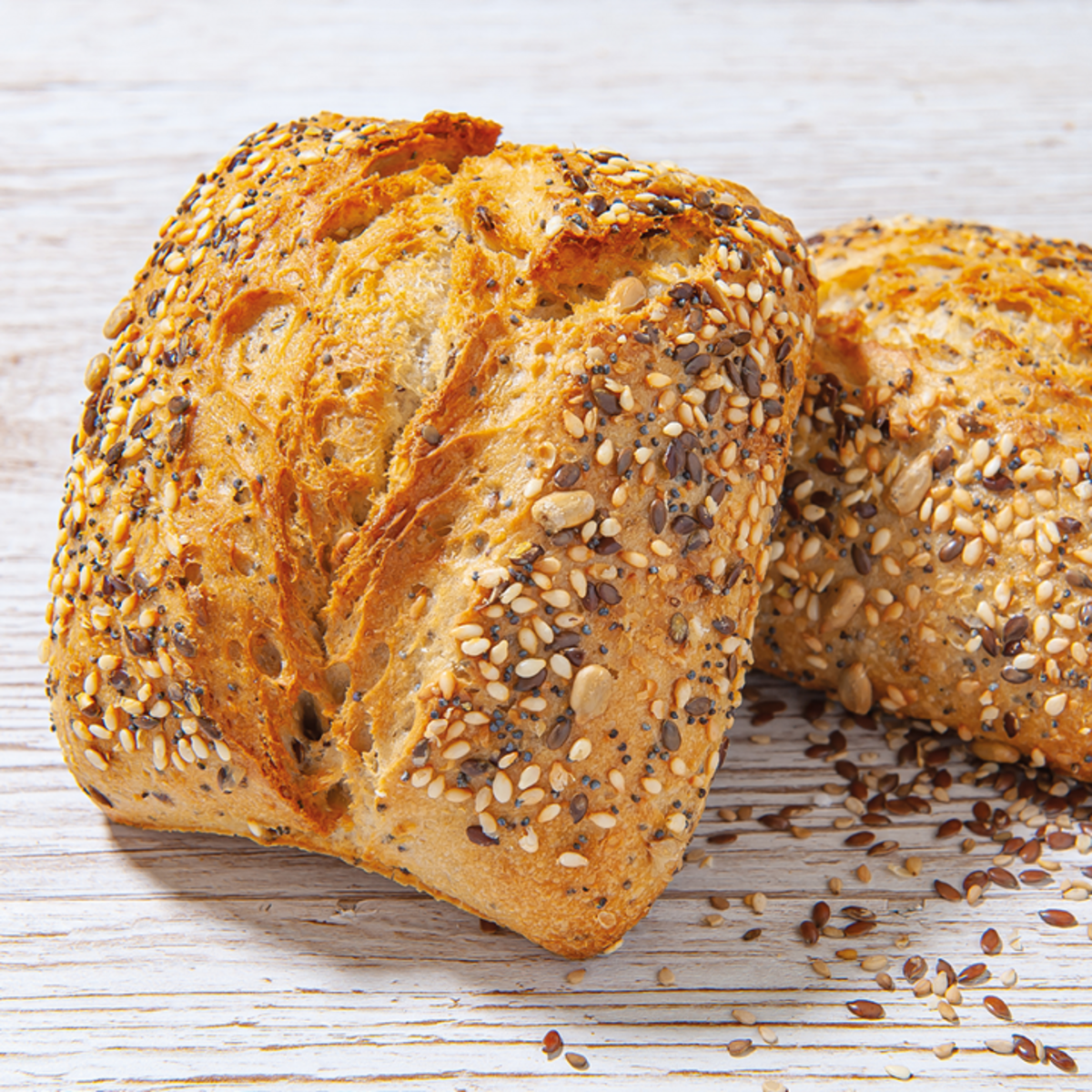 Zwei Brötchen mit einer knusprigen Kruste, bestreut mit verschiedenen Samen, liegen auf einer hellen Holzoberfläche. Einige Samen sind um die Brötchen verstreut.