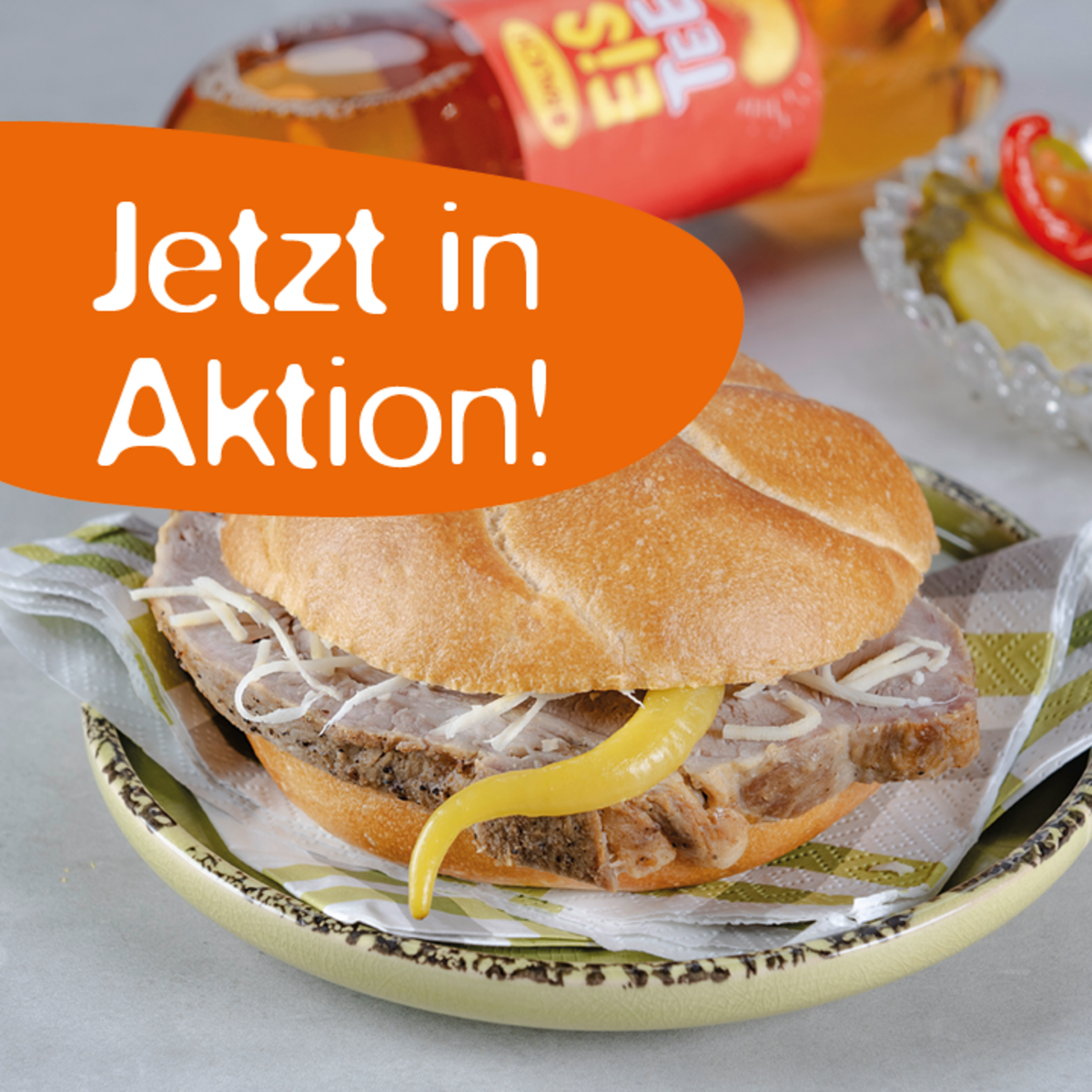 Ein Sandwich mit Fleisch, Zwiebeln und einer gelben Paprika auf einem Teller. Im Hintergrund steht eine Flasche Eistee.